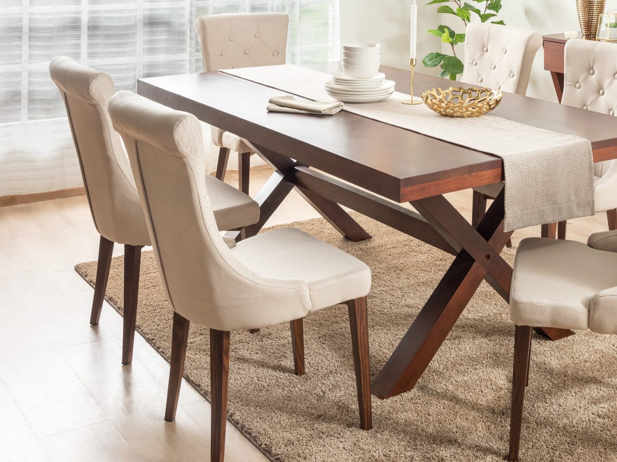 A dark wood dining table with a cross-braced base, set with white dishes and surrounded by cream upholstered chairs.
